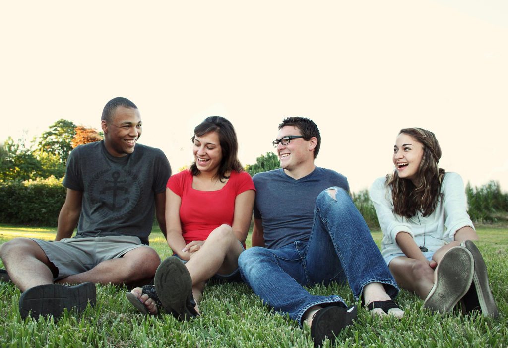 A group of young adults smiling while sitting on grass in a sunny park, enjoying friendship and leisure.