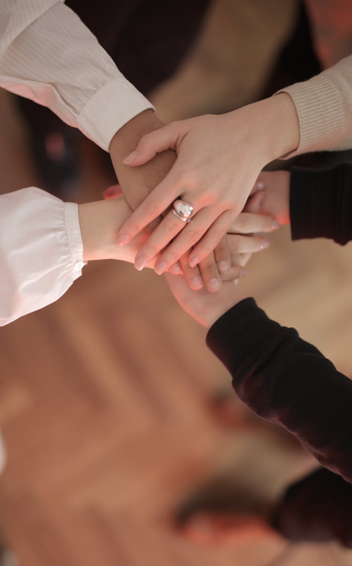 about-01 Top view of faceless friends in different clothes stacking hands together while standing on wooden floor indoor on sunny day
